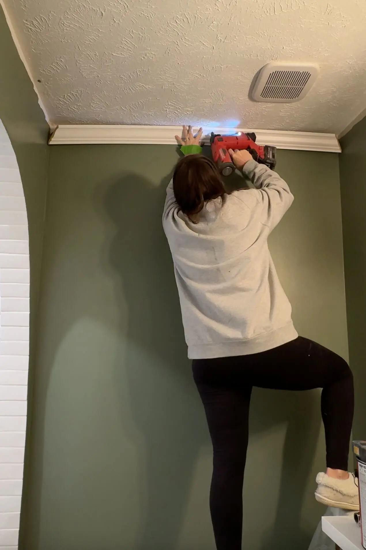 Person installing white crown molding on a green bathroom wall using a nail gun, highlighting a hands-on step in a DIY home improvement project.